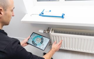 Osoba trzymająca tablet reguluje temperaturę kaloryfera, co podnosi ciepło we wspólnocie mieszkaniowej.
