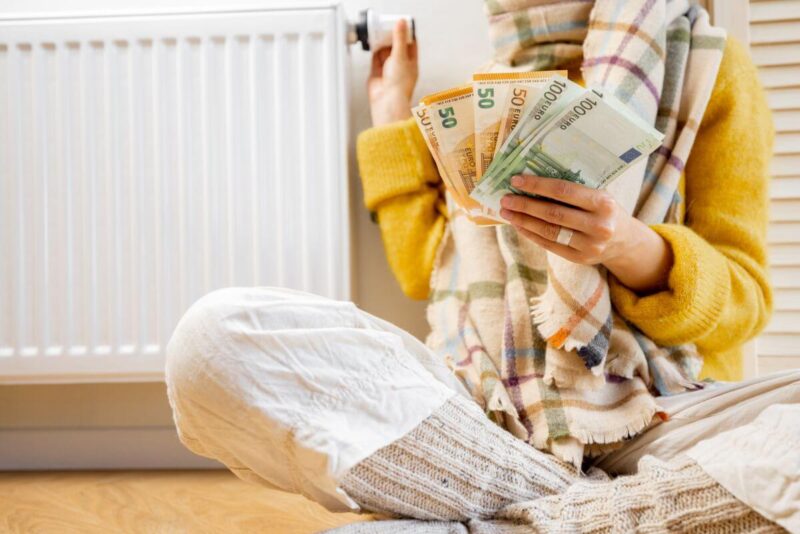 Osoba przy grzejniku trzyma gotówkę, sugerując dopłatę za ogrzewanie.