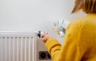 Osoba w żółtym swetrze trzyma pieniądze przy kaloryferze, wskazując na wysokie rachunki zaciepło.